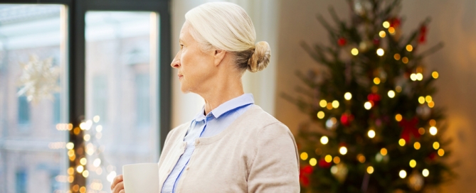 senior woman with cup of tea or coffee looking through window at home over christmas tree lights background