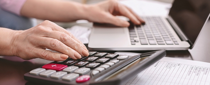 Senior Woman Calculating Taxes At Home, Using Calculator And Laptop tax deduction