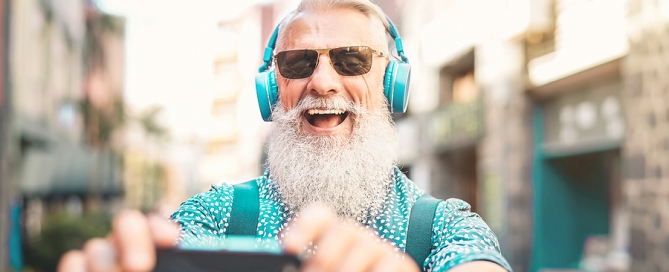Senior bearded man taking selfie with mobile phone while listening favorite playlist with headphones younger seniors subjective age