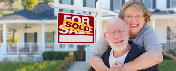 Senior Adult Couple in Front of Sold Home For Sale sign; selling your home
