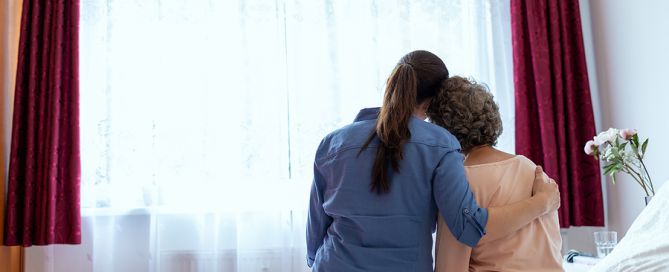 dementia caregiving; woman hugging elderly woman by window