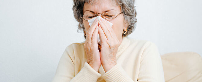 Portrait of sick senior woman sneezing, coughing into napkin while sitting on couch CCRC tripledemic