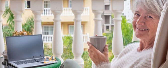 Choosing a CCRC Residential Unit Smiling senior woman relaxing on home balcony while using laptop drinking coffee