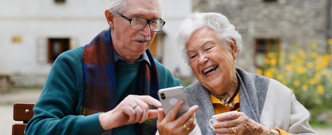 Happy senior couple in cafe having coffee and looking at smartphone; memory care assisted living